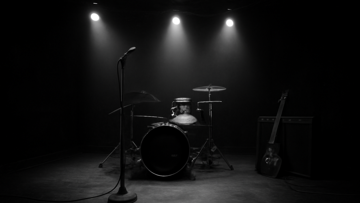 Panggung klub rock kecil dengan mikrofon, drum, dan gitar bersandar, suasana sunyi sebelum pertunjukan